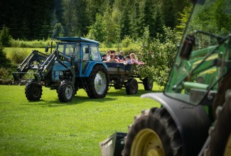 Sinine traktor, mille järelkärus istuvad täiskasvanud ja lapsed, taustal mets.